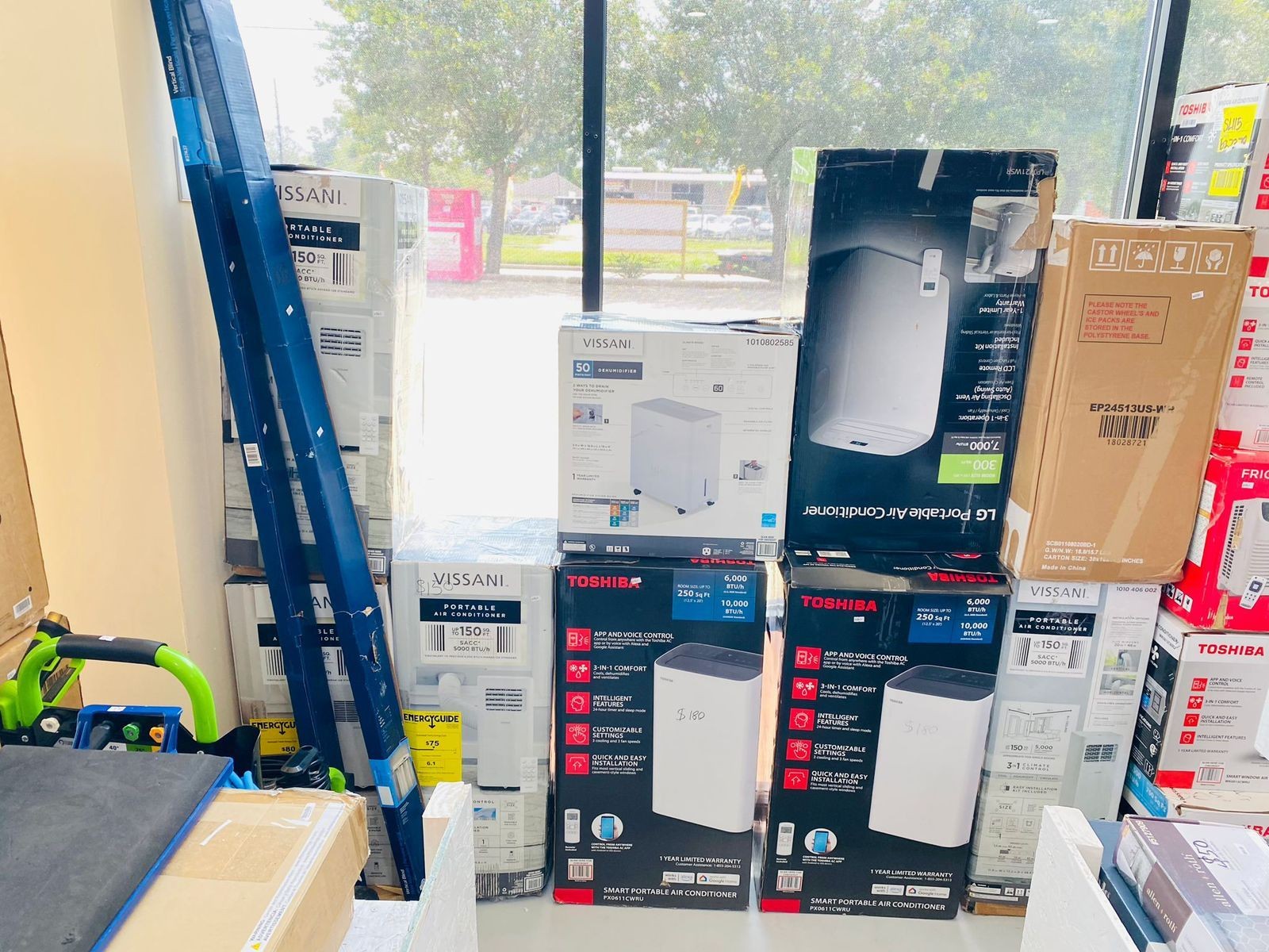 A store display featuring stacked boxes of portable air conditioners and other appliances near a large window.